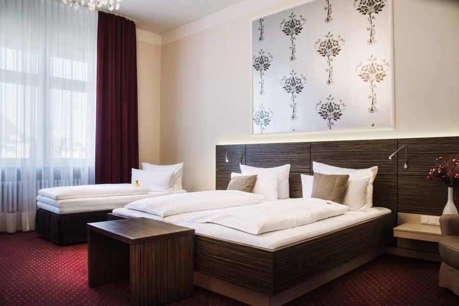 Ein Deluxe‑Doppelzimmer mit großem Bett, zusätzlichem Einzelbett, floralem Wandmotiv und Fenster mit Vorhängen im Hotel Prinzregent.