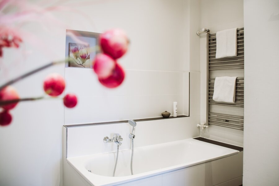 Ein Deluxe‑Bad mit weißer Badewanne, Handbrause, Handtuchwärmer und Blumen im Vordergrund im Hotel Prinzregent.