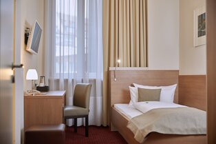 Ein kompaktes Hotelzimmer mit Einzelbett, Schreibtisch mit Lampe und Wasserkocher, Wand‑TV und Fenster mit Vorhängen im Hotel Prinzregent.