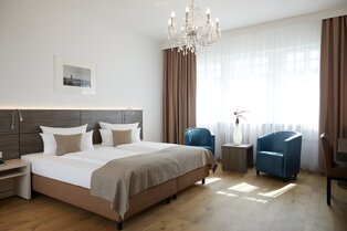 Elegantes Doppelzimmer im Hotel Prinzregent mit Kronleuchter, zwei blauen Sesseln und großem Doppelbett.