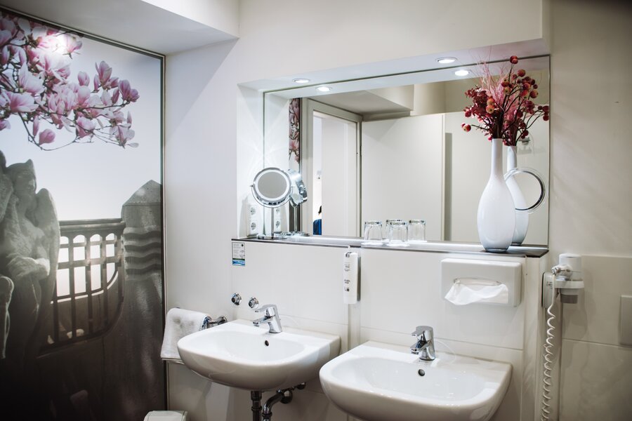 Ein Deluxe‑Bad mit zwei Waschbecken, großem Spiegel, Föhn und dekorativer Blumen­nische im Hotel Prinzregent.