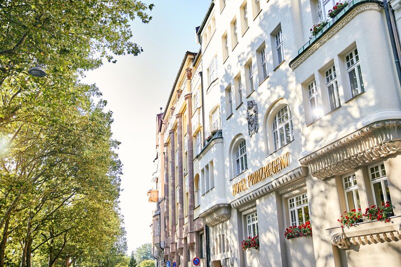 Außenansicht der historischen Fassade des Hotel Prinzregent mit blühendem Blumenschmuck und angrenzender Allee.