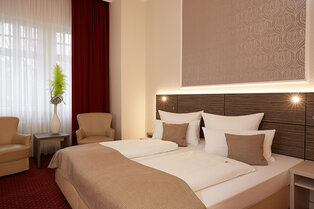 Ein modernes Doppelzimmer mit großem Bett, zwei Sesseln, kleinem Tisch mit Vase und bodenlangen Vorhängen im Hotel Prinzregent.