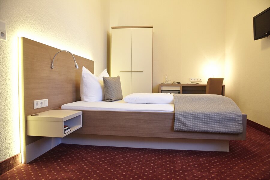Ein schlichtes Einzelzimmer mit Bett, Schreibtisch, Kleiderschrank und rotem Teppich im Hotel Prinzregent.