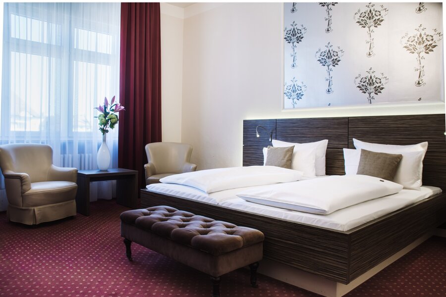 Ein Deluxe‑Doppelzimmer mit großem Bett, zwei Sesseln, kleinem Tisch mit Blumen und rotem Teppich im Hotel Prinzregent.