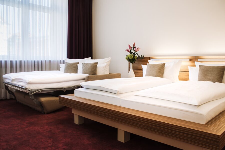 Ein Deluxe‑Doppelzimmer mit großem Bett, zusätzlichem Schlafsofa und Blumenarrangement vor dem Fenster im Hotel Prinzregent.