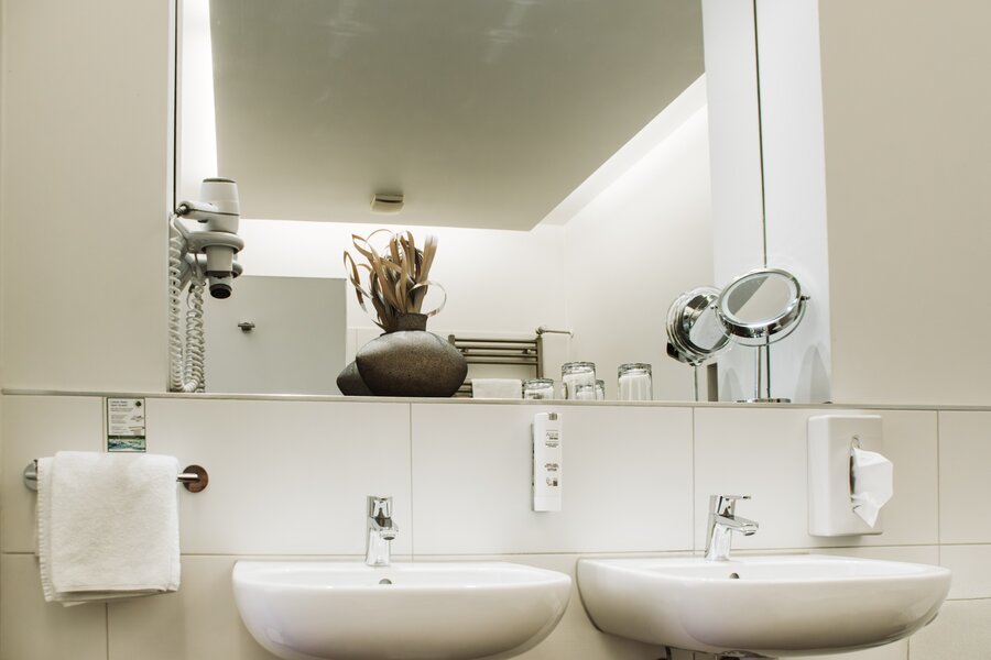 Ein Deluxe‑Bad mit zwei Waschbecken, großem Spiegel, Föhn, Gläsern und dunkler Vase mit Trockenpflanzen im Hotel Prinzregent.