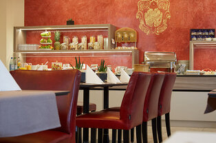 Ein Frühstücksraum mit weiß gedeckten Tischen und rotem Lederstühlen, im Hintergrund ein Buffet mit Getränkespendern, Müsligläsern, Obst und Warmhaltebehälter im Hotel Prinzregent.
