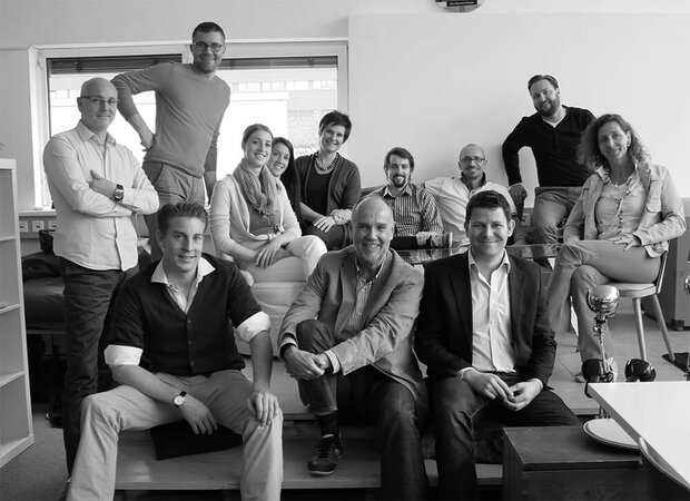    Ein schwarz‑weißes Gruppenfoto von zwölf Personen in einem modernen Büro, sitzend und stehend vor Fenstern.