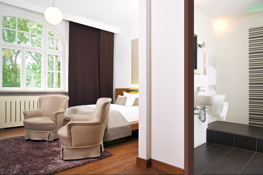 Ein Superior‑Doppelzimmer mit großem Bett, zwei Sesseln und Blick durchs Fenster auf Bäume im Hotel Prinzregent.