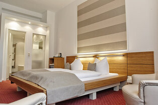 Modern eingerichtetes Deluxe-Doppelzimmer im Hotel Prinzregent mit Holzbett, dezentem Wanddekor und Sessel.