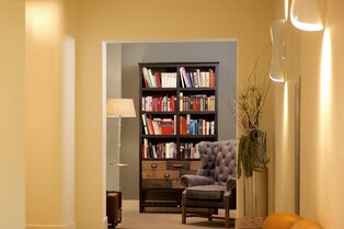 Eine gemütliche Leseecke mit grauem Sessel, Stehlampe, Bücherregal und hoher Vase im Hotel Prinzregent.