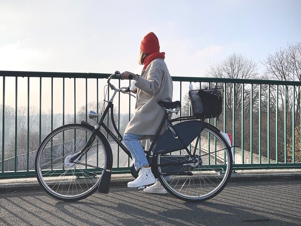Eine Frau in einem beigen Mantel und orangefarbener Mütze sitzt auf einem schwarzen Fahrrad auf einer Brücke.