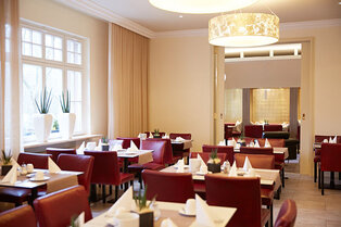 Ein heller Frühstücksraum mit weiß gedeckten Tischen, roten Stühlen, großen runden Deckenleuchten und Fenstern im Hotel Prinzregent.