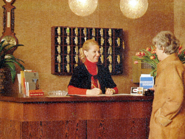    Die Hotelrezeption mit Mitarbeitenden, Schlüsselwand, Prospekten und zwei runden Lampen im Hotel Prinzregent.