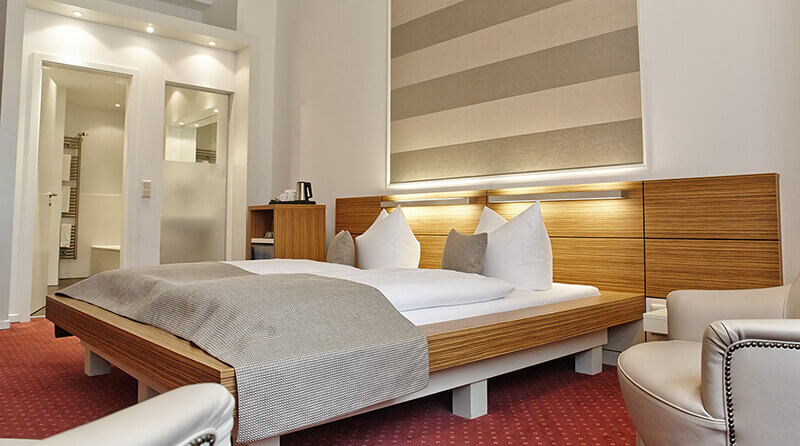 Ein Deluxe‑Doppelzimmer mit großem Bett, warmem Wandlicht, rotem Teppich und Blick ins Bad im Hotel Prinzregent.
